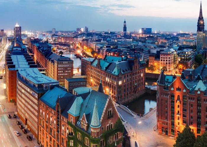 Abendliche Stadtansicht der Speicherstadt in Hamburg, mit ihren historischen Backsteingebäuden entlang der Kanäle, beleuchtet von warmem, gelben Licht; im Hintergrund sind die Kirchtürme und moderne Gebäude der Stadt erkennbar.