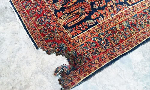 Ecke eines Teppichs mit blumigem orientalischem Muster, teilweise beschädigt und auf einem hellen Boden liegend.