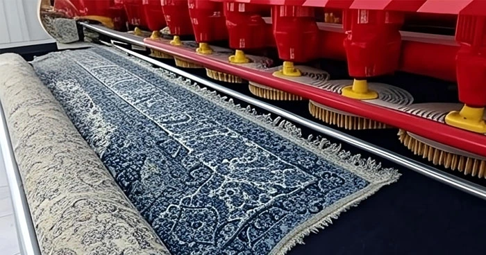 Ein blauer Teppich mit komplizierten Mustern wird durch eine industrielle Teppichreinigungsmaschine gereinigt, die aus mehreren rot-gelben Bürsten besteht.