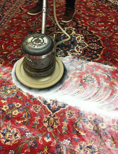 Ein großer, runder Teppichreiniger wird verwendet, um einen roten, gemusterten Teppich zu reinigen, wobei Seifenschaum sichtbar ist.