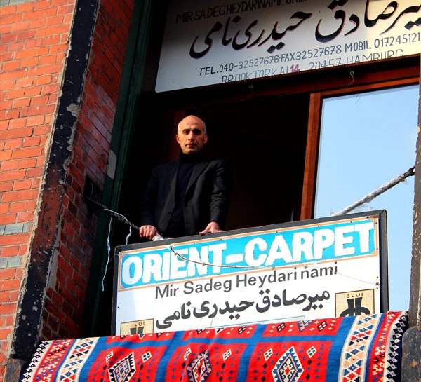 Ein Mann steht auf einem Balkon vor einem Geschäftsschild mit der Aufschrift "Orient-Carpet" und einem Teppich im Vordergrund.
