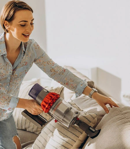 Eine Frau in einem hellblauen Hemd saugt mit einem tragbaren Staubsauger ein Sofa ab.