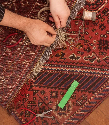 Eine Person repariert die Fransen eines roten, gemusterten Teppichs mit Garn und Nadeln, während Werkzeuge wie eine Schere und ein Maßband daneben liegen.
