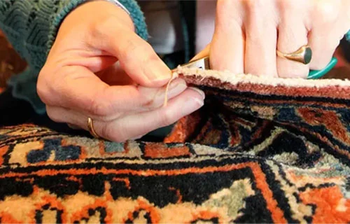 Eine Person repariert einen orientalischen Teppich mit Nadel und Faden, wobei nur die Hände zu sehen sind.