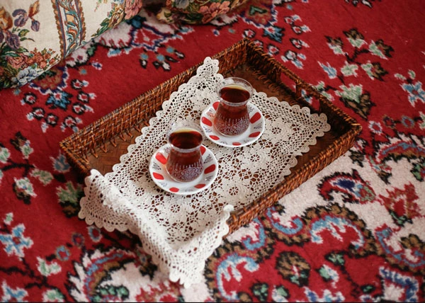 Zwei Gläser mit türkischem Tee auf Untertassen stehen auf einem geflochtenen Tablett mit einem Spitzenläufer, auf einem roten Teppich mit floralem Muster platziert.